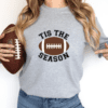 woman wearing a gray crewneck sweatshirt with the phrase "Tis the Season" and a football on shirt.