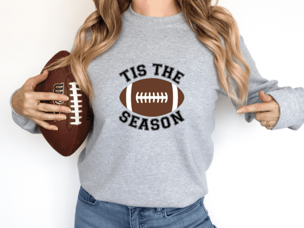 woman wearing a gray crewneck sweatshirt with the phrase "Tis the Season" and a football on shirt.