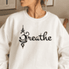 woman wearing a white sweatshirt with the word "Breathe" on the front in black. The "B" in Breathe is the symbol for Namaste instead of an actual letter B.