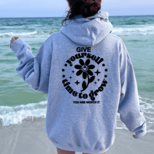 woman wearing a gray hoodie walking near ocean. hoodie reads "give yourself time to grow. you deserve it"