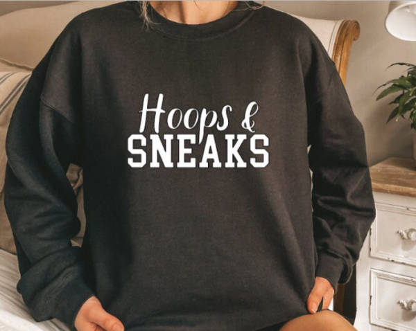 Woman wearing a black crewneck sweatshit with the phrase "Hoops and Sneaks" written in white across the chest.