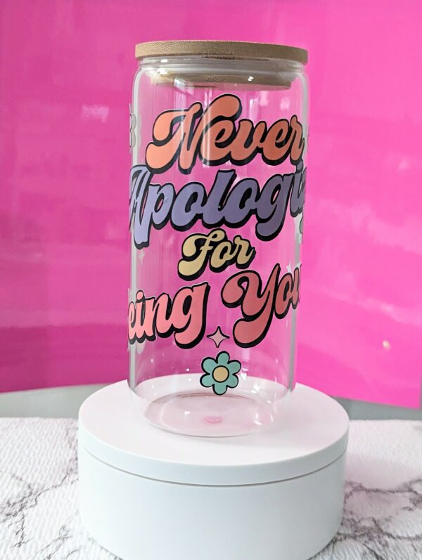 Transparent glass can with bamboo lid with the phrase "Never Apologize for Being You" written in colorful letters. Colorful nostalgic daisy decorations on the sides.