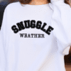 Woman wearing a white sweatshirt with the phrase "Snuggle Weather" written in black college letters.