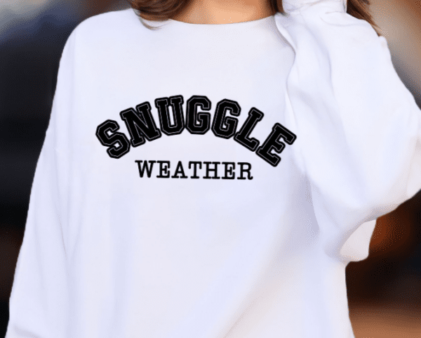 Woman wearing a white sweatshirt with the phrase "Snuggle Weather" written in black college letters.
