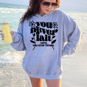 Woman on the beach wearing a gray hoodie with the phrase "You never fail until you stop trying" holding an insulated tumbler.