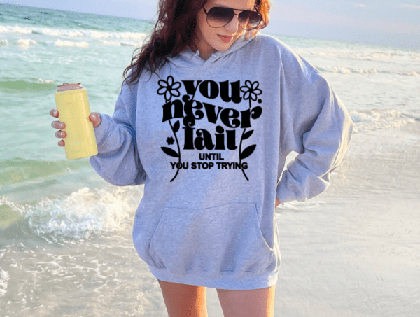 Woman on the beach wearing a gray hoodie with the phrase "You never fail until you stop trying" holding an insulated tumbler.
