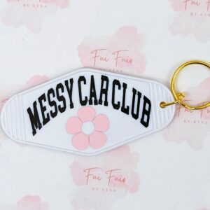 White motel keychain with gold tone ring and jump ring. The phrase "Messy Car Club" in black and a pink flower underneath.