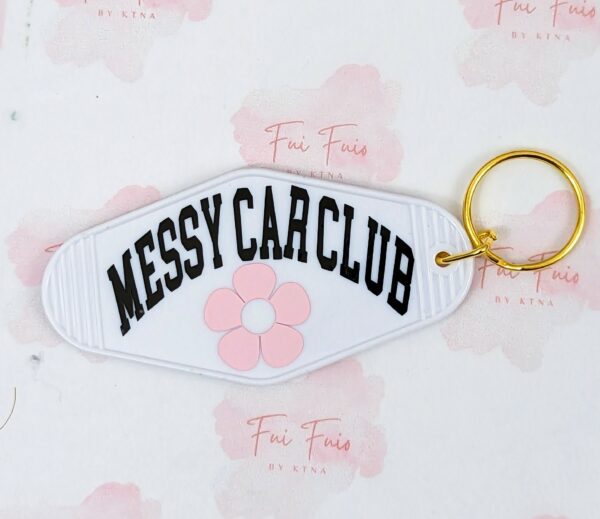 White motel keychain with gold tone ring and jump ring. The phrase "Messy Car Club" in black and a pink flower underneath.