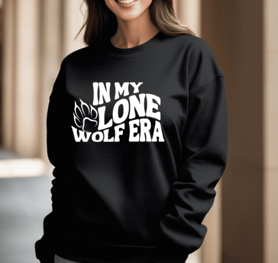 woman wearing a black crewneck sweatshirt with the phrase "in my lone wolf era" written in a retro wavy font with a wolf paw print next to the word "lone"