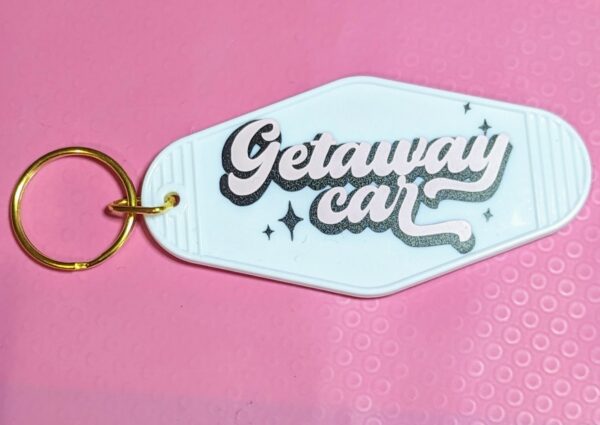 White motel keychain with gold keying. Phrase "Getaway Car" written in pink with black shadow. Retro font.
