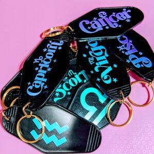 Pile of black motel keychains with gold key rings and astrological signs written on one side. The other side has the astrological side symbol. Holographic.