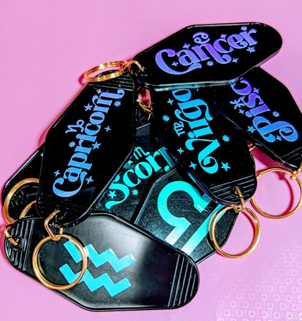 Pile of black motel keychains with gold key rings and astrological signs written on one side. The other side has the astrological side symbol. Holographic.