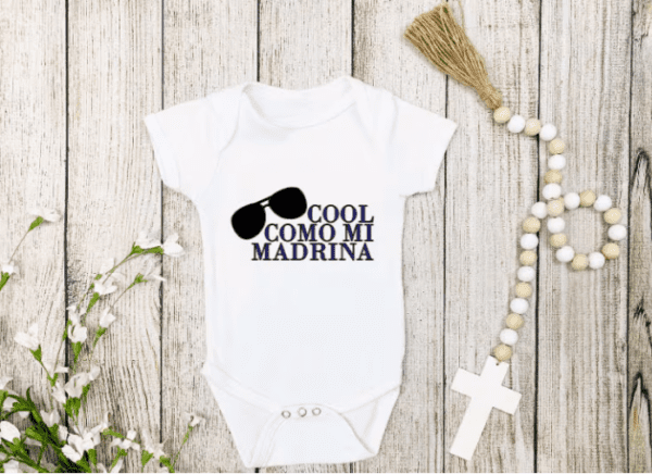 White one-piece baby bodysuit with the phrase "Cool Like Mi Madrina" with the silhouette of sunglasses diagonally on the side.