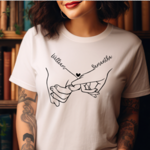 woman wearing a white t-shirt with couples hands holding each others pinkys. The wrist above the pinky has the couples first names connecting to the hand.