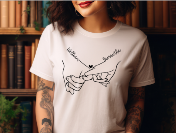 woman wearing a white t-shirt with couples hands holding each others pinkys. The wrist above the pinky has the couples first names connecting to the hand.