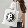 Woman with white tote bag on her shoulder. The bag has the yin and yang symbol with hearts where the circles normally are. The quote circling the yin and yang reads, "Find the balance between heart and mind"