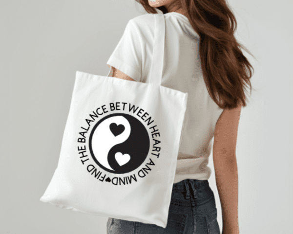 Woman with white tote bag on her shoulder. The bag has the yin and yang symbol with hearts where the circles normally are. The quote circling the yin and yang reads, "Find the balance between heart and mind"