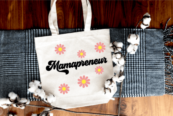 white tote bag with the word "mamapreneur" written on the front in a retro font. Individual daisy like flowers are above and below the word.