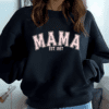 Woman wearing a black crewneck sweatshirt with the words MAMA EST with the year of choice. The letters are a light pink.