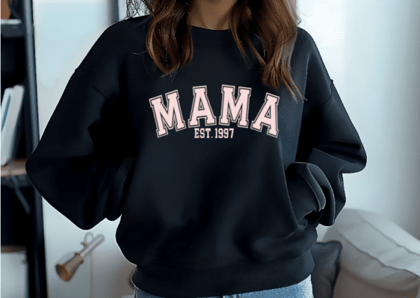 Woman wearing a black crewneck sweatshirt with the words MAMA EST with the year of choice. The letters are a light pink.