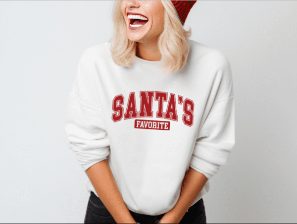 Woman wearing a white crewneck sweatshirt with the phrase "Santa's Favorite" written in red collegiate font.