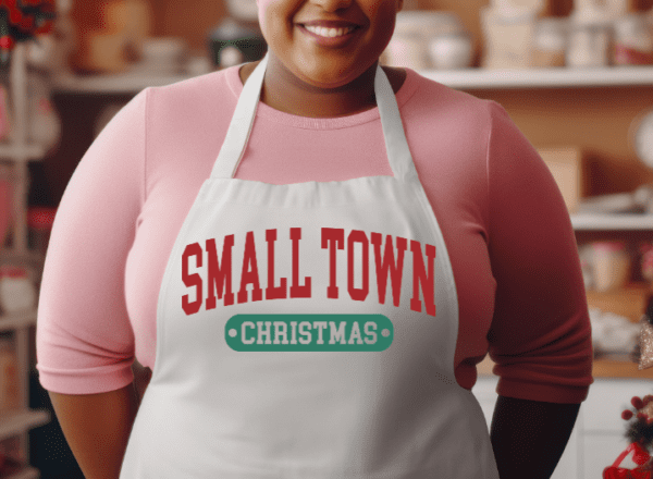Woman wearing a white apron with the phrase Small Town in red and Christmas underneath in green.