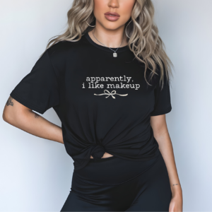 Person wearing a black t-shirt with white lowercase text that reads “apparently, i like makeup” and a small bow graphic. Shirt is knotted at the waist. Model has long wavy hair, visible tattoos, and wears black pants. Background is light gray.