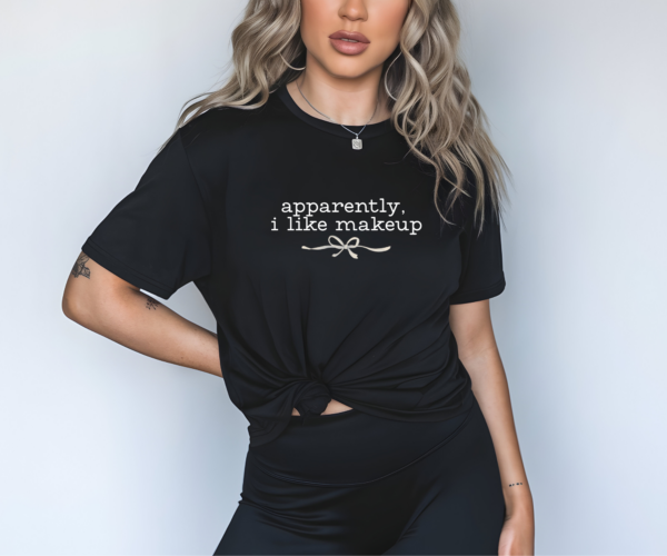 Person wearing a black t-shirt with white lowercase text that reads “apparently, i like makeup” and a small bow graphic. Shirt is knotted at the waist. Model has long wavy hair, visible tattoos, and wears black pants. Background is light gray.