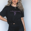 Person wearing a black t-shirt with white lowercase text that reads “i do all the pretty girls makeup” and a small bow graphic. Shirt is knotted at the waist. Model has long wavy blonde hair, visible tattoos, and wears black pants. Background is neutral and softly lit.
