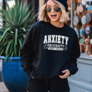 Person wearing a black unisex sweatshirt that says “Anxiety University Honors Program” while standing outdoors near a storefront.