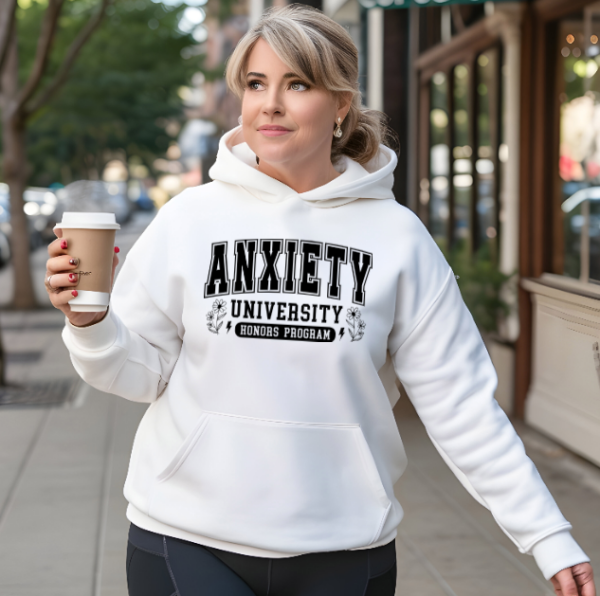 Woman walking on the sidewalk by a storefront holding a coffee cup. The hoodie reads "Anxiety University Honors Program"