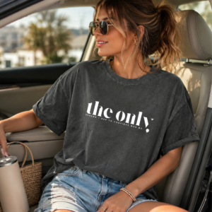 Person wearing a dark gray oversized T‑shirt that reads “the only person I had to convince was me,” sitting in a car holding a tumbler.